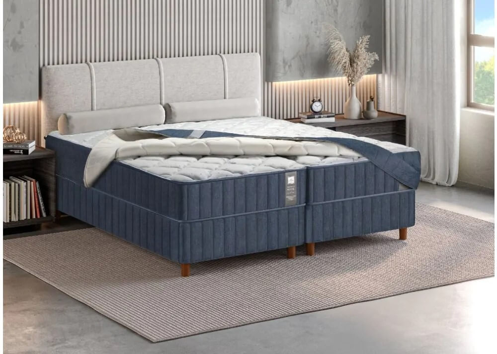 Vista em perspectiva de uma cama de casal de alto padrão composta por dois box articulados revestidos em tecido azul marinho com costura vertical. Sobre os colchões, há uma capa protetora acolchoada com acabamento em matelassê branco na parte superior e laterais em azul, que está parcialmente dobrada, revelando a textura interna do colchão. A cabeceira é estofada em tom bege claro e o ambiente possui decoração moderna e minimalista.