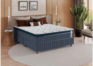 Cama de casal completa em um quarto com painéis de madeira. A base box e as laterais do colchão são revestidas em tecido azul texturizado. O topo apresenta uma capa de colchão branca e volumosa com costura profunda em gomos (estilo capitonê), conferindo um aspecto de maciez e luxo. Almofadas em tons de marrom e preto decoram a cabeceira de palha natural.