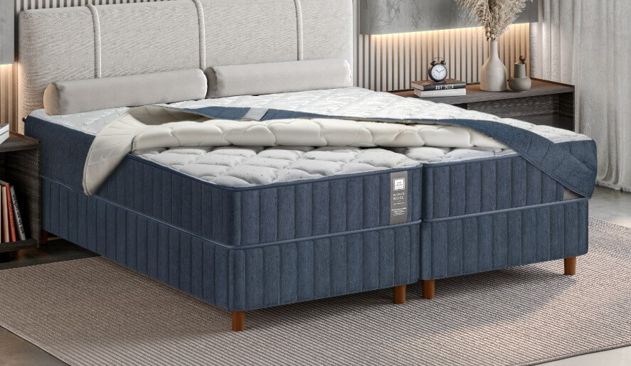 Vista em perspectiva de uma cama de casal de alto padrão composta por dois box articulados revestidos em tecido azul marinho com costura vertical. Sobre os colchões, há uma capa protetora acolchoada com acabamento em matelassê branco na parte superior e laterais em azul, que está parcialmente dobrada, revelando a textura interna do colchão. A cabeceira é estofada em tom bege claro e o ambiente possui decoração moderna e minimalista.
