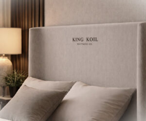 Close-up da cabeceira estofada que compõe a Cama Elegance, mostrando a logomarca King Koil bordada centralizada. O tecido premium complementa o design funcional da base Box Baú.